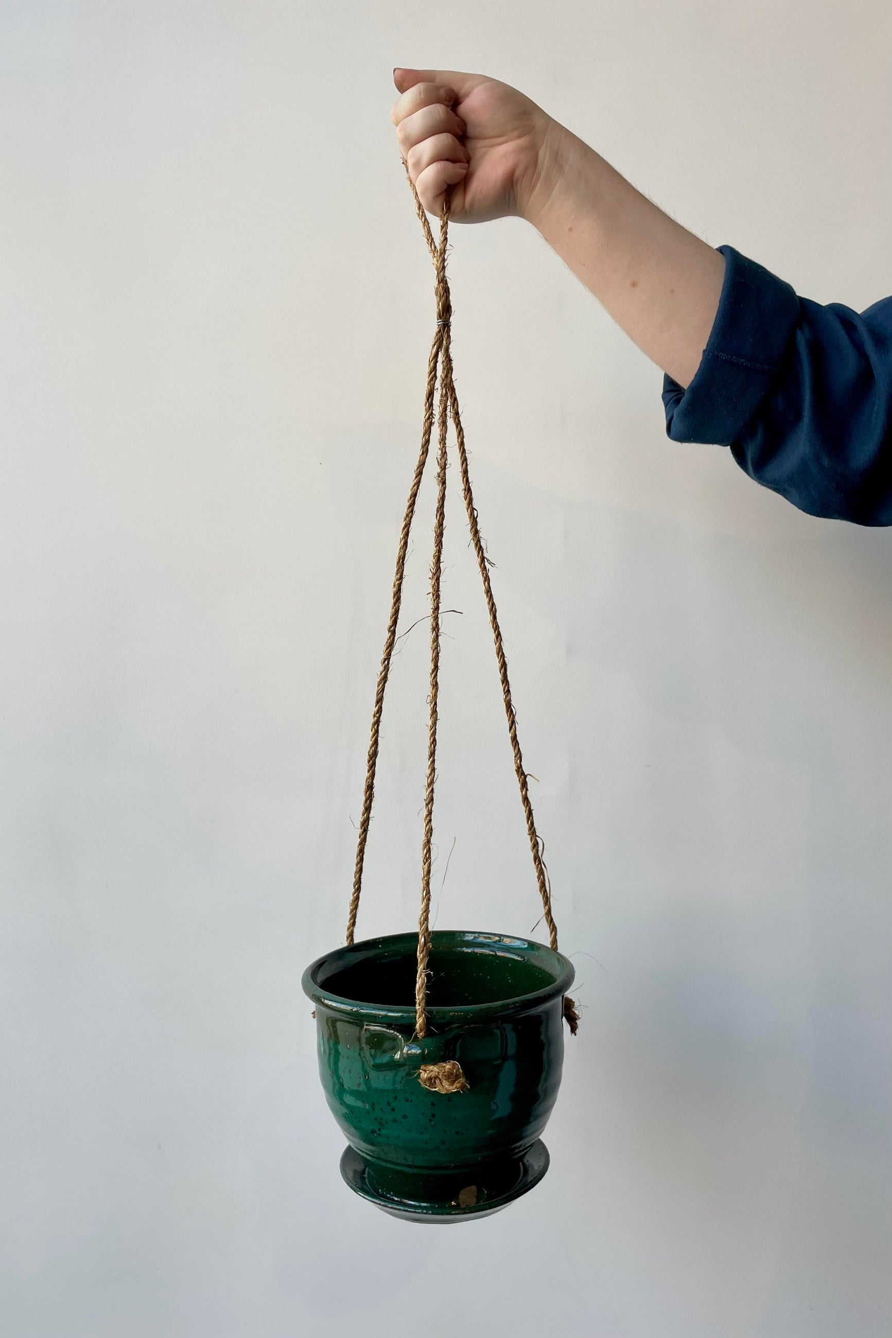 Six inch clay planter with drainage hole and attached tray in a jade green glaze flecked with silver specks against a white background. The planter has a jute cord attached at three points to hang. A hand is holding this planter against a white wall. ©Sprout Home