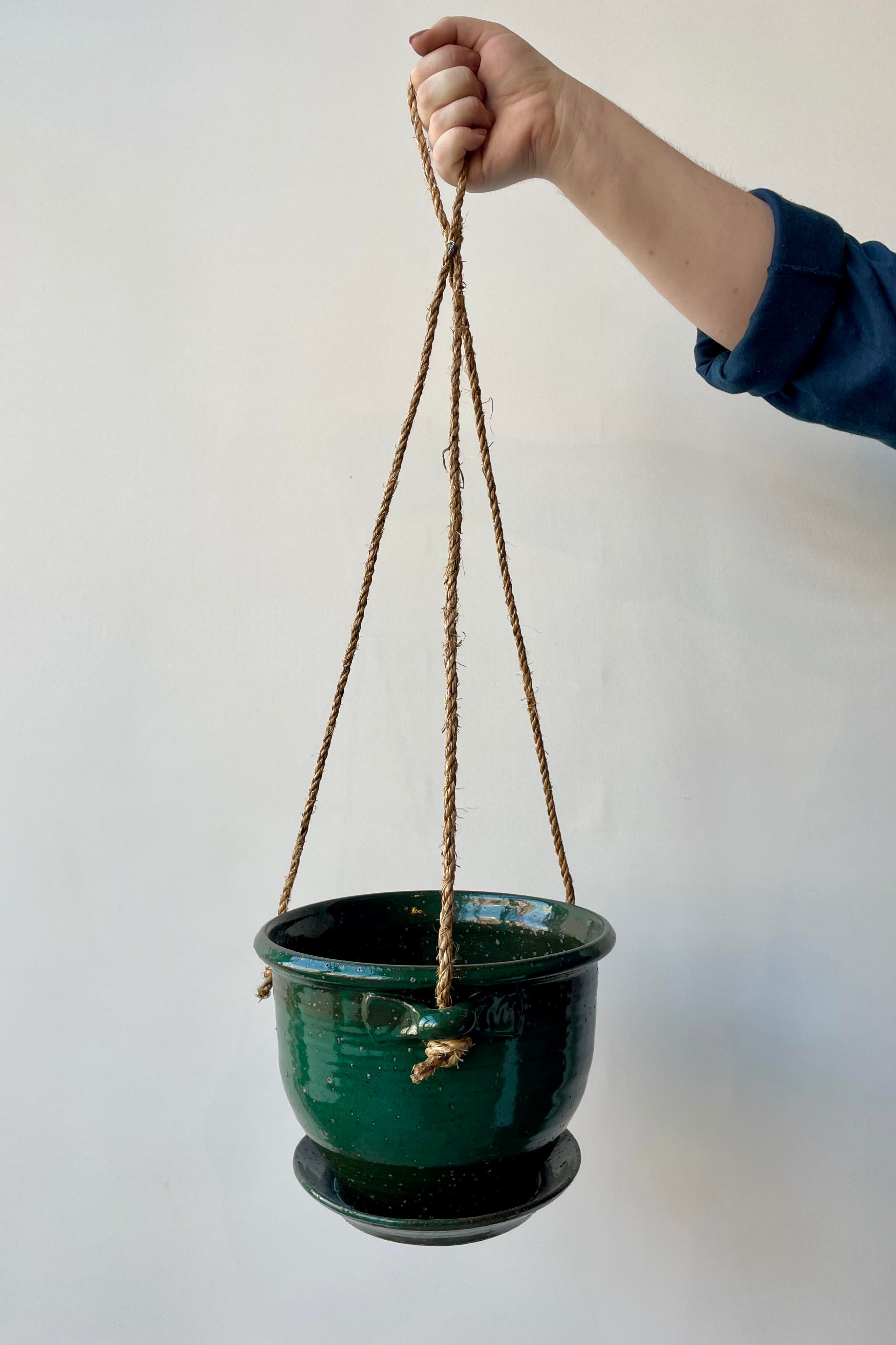 Eight inch clay planter with drainage hole and attached tray in a jade green glaze flecked with silver specks against a white background. The planter has a jute cord attached at three points to hang. A hand is holding this planter against a white wall.