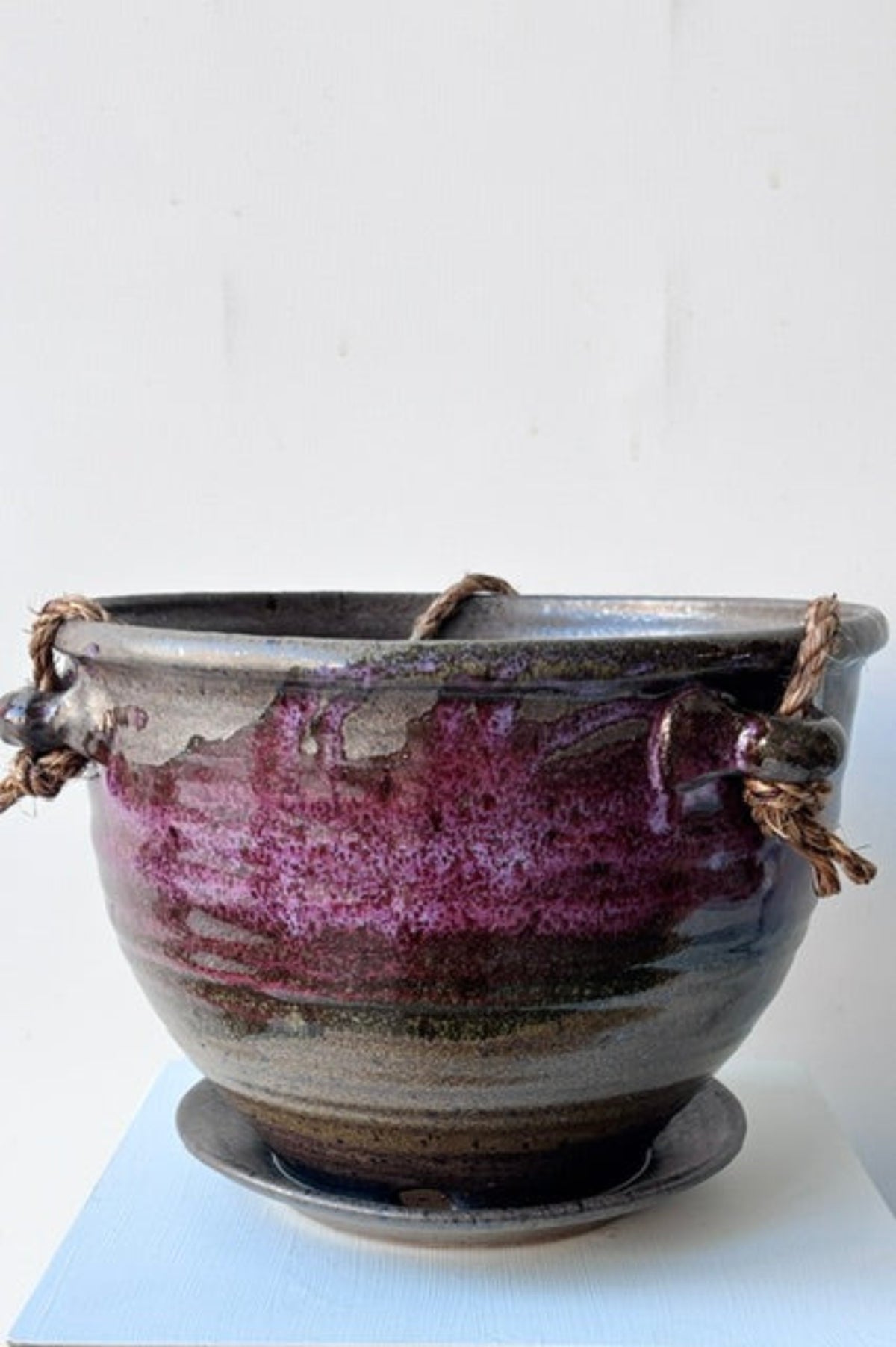 A handmade ceramic planter pot with a dark brown and purple glaze, featuring two rope handles on the sides and a matching saucer, set against a plain white background.