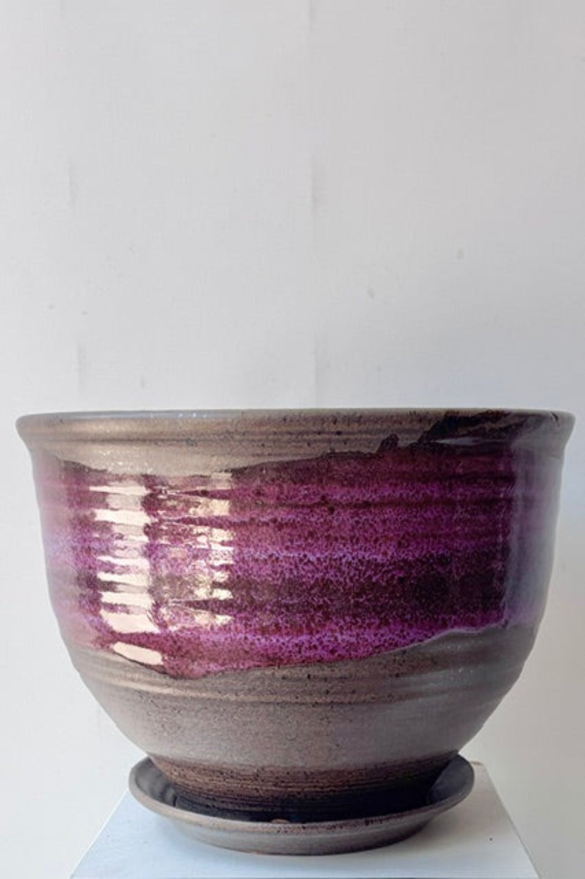 A handmade ceramic planter pot with a matching saucer, featuring a glossy purple and brown glaze, displayed on a white pedestal against a white wall.
