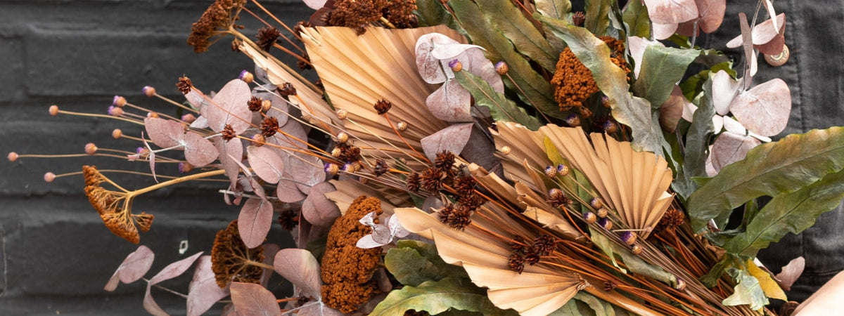 Preserved floral containing brown Yarros, natural palm leaves, rust eucalyptus, and green fern leaves against a black wall ©Sprout Home