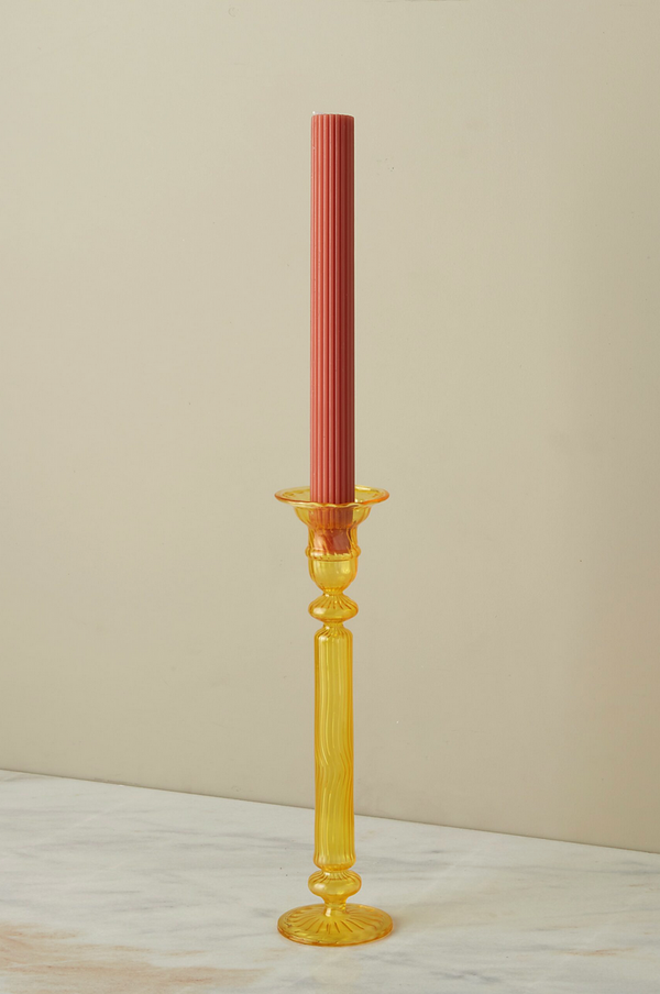 A Nahlia Daffodil colored glass candlestick holder with a terracotta colored taper candle unlit. 