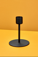 The Kent Tapered black Candlestick holder medium against a yellow wall and table. 