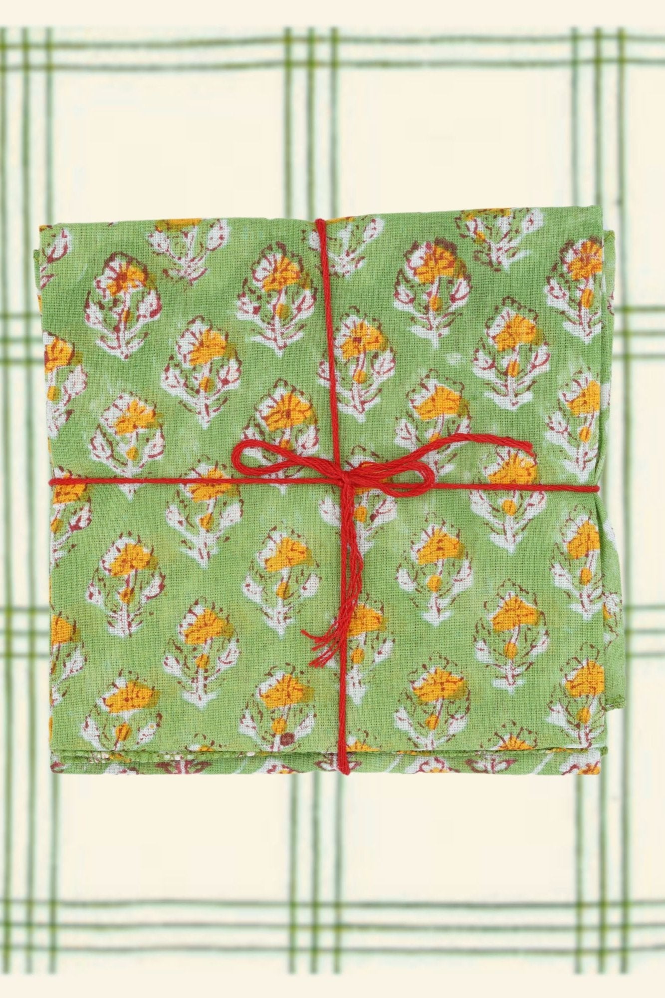 Suzette Achiel Tilleul set of four napkins folded and tied with a red string on a green and white plaid background. 