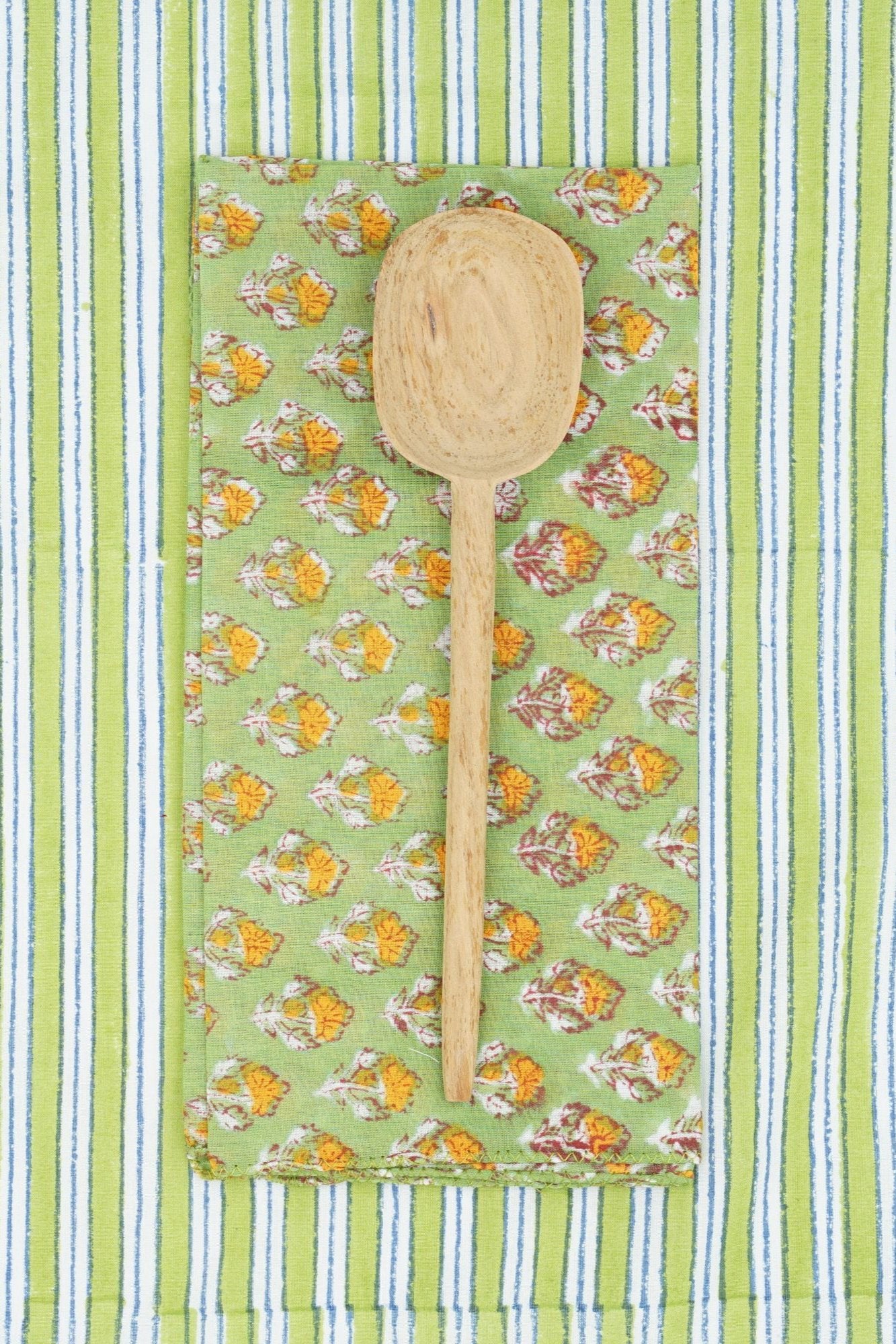 Suzette Achiel Tilleul napkin folded in to a rectangle and a wood spoon on top with a striped background. 