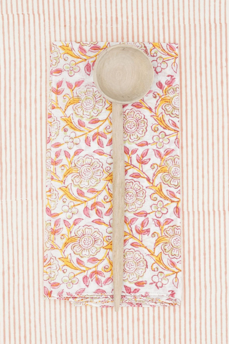 Suzette Frangipanier Jaipur napkin folded in to a rectangle, wood spoon on top and on a striped background. 