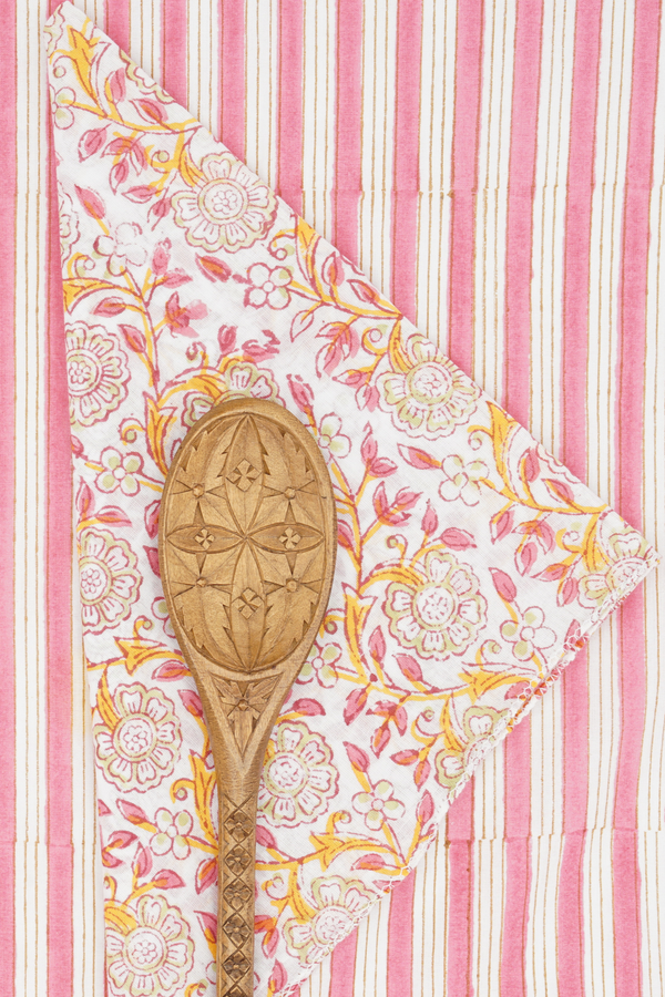 Suzette Frangipanier Jaipur napkin folded in to a triangle, wood spoon on top and on a pink and white striped background. 