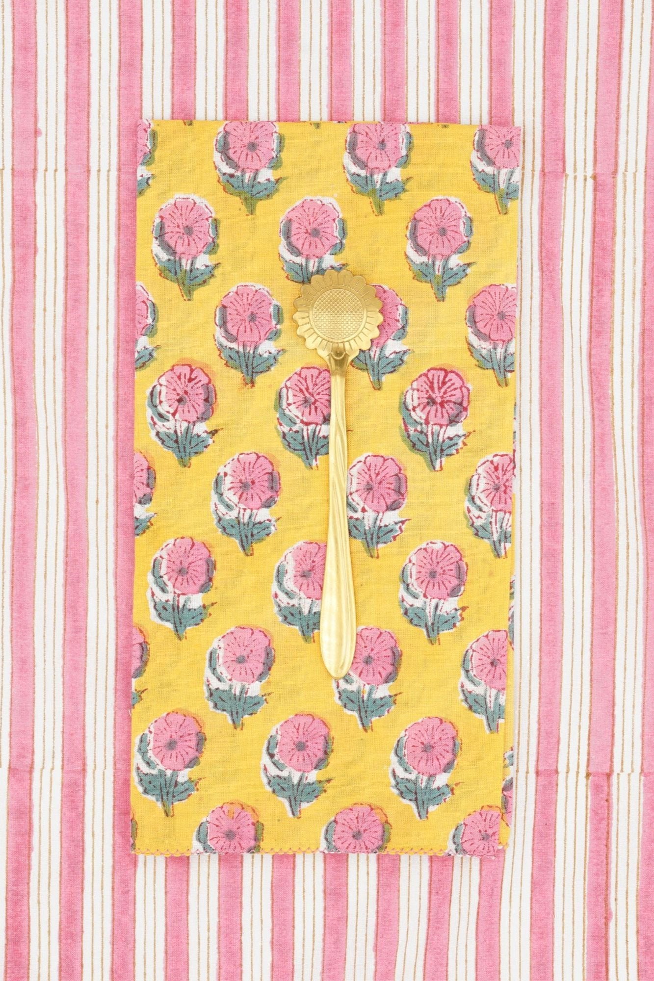 Suzette Cotton Napkins Helenium Lemon folded into a rectangle, grass utensil on top and a pink and white striped background. 