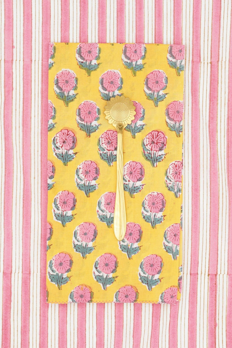 Suzette Cotton Napkins Helenium Lemon folded into a rectangle, grass utensil on top and a pink and white striped background. 