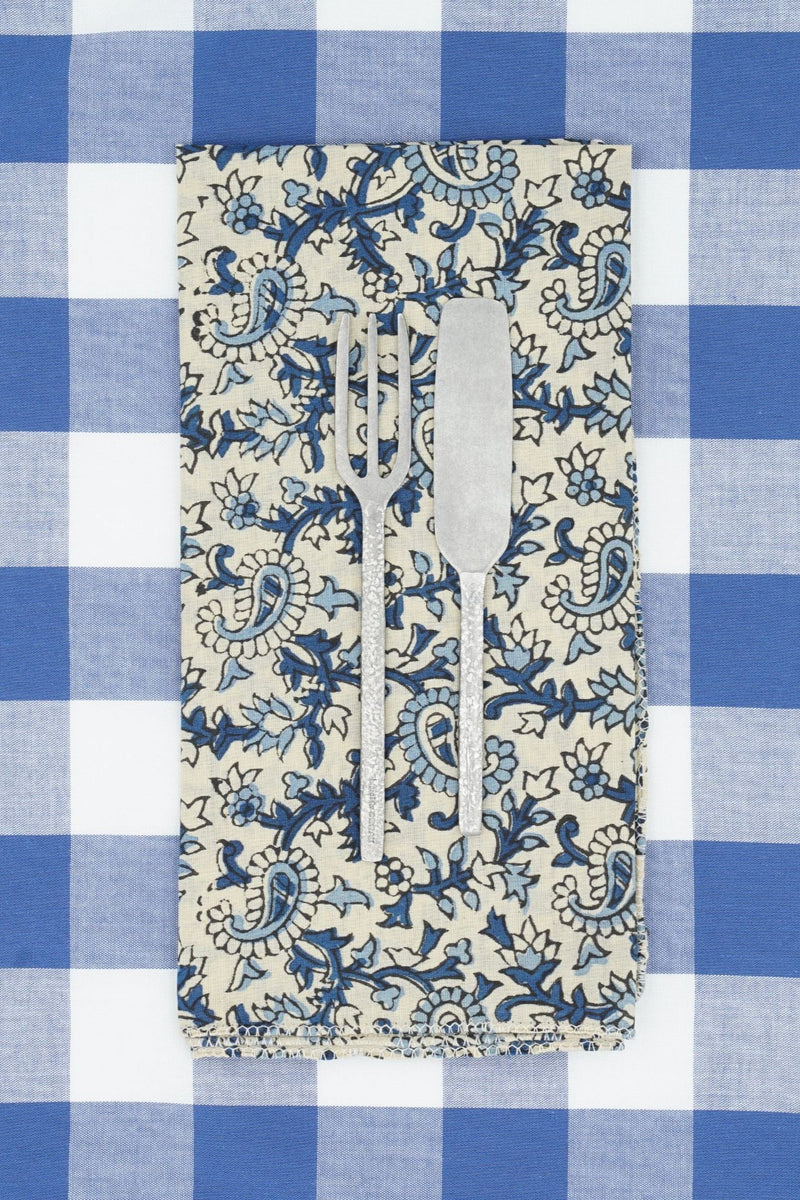 Suzette Cotton Napkin Pourpier Cobalt folded in to a rectangle with silver flatware on top and sitting on a blue and white checkered textile. 