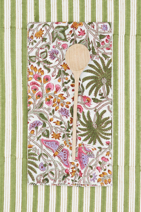 Suzette Cotton Napkin Primavera Cyprès folded in a rectangle, wood spoon on top and sitting on a green and white linen. 