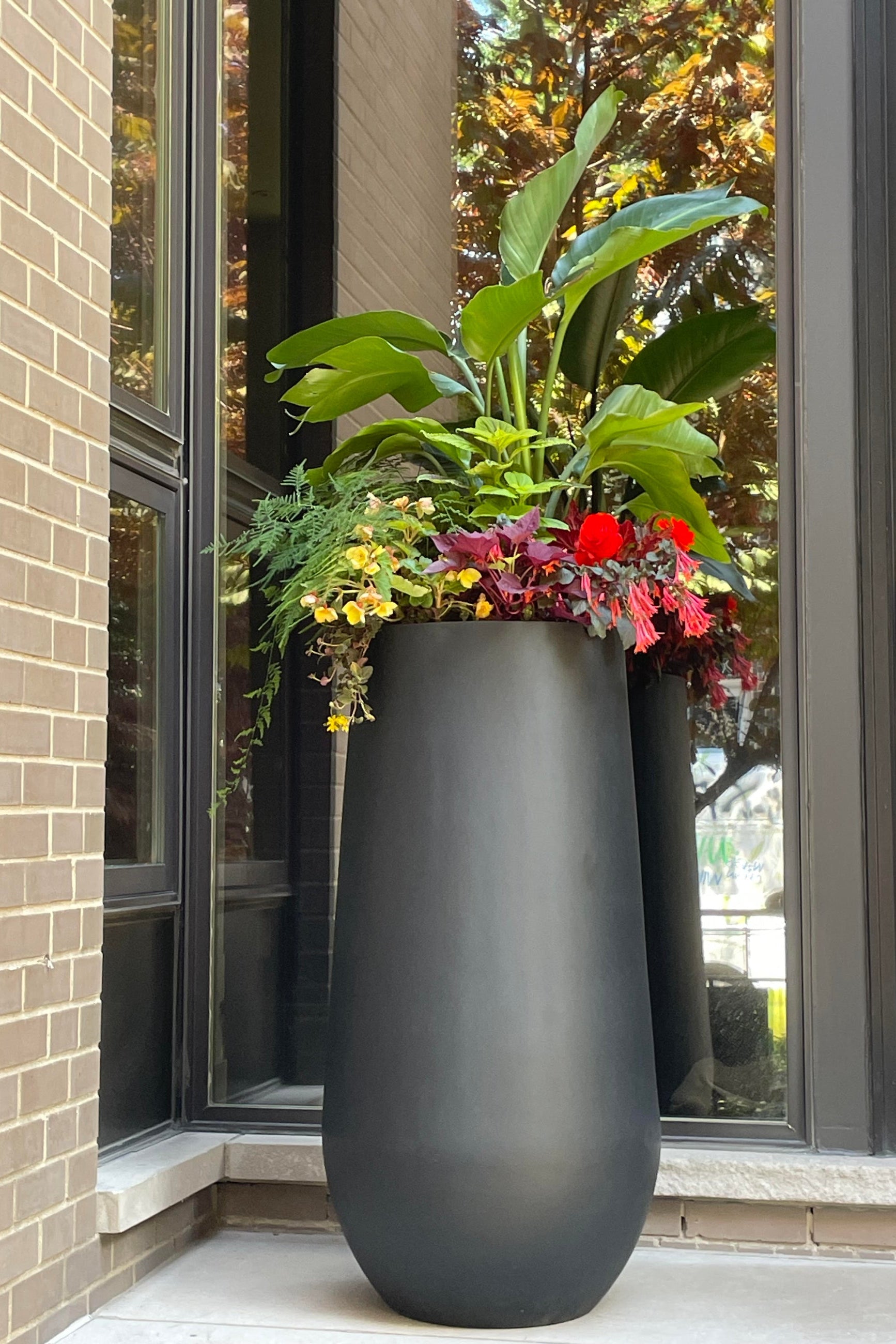 Photo of tall slender black pot in a brick seting with colorful tropical flowers ©Sprout Home