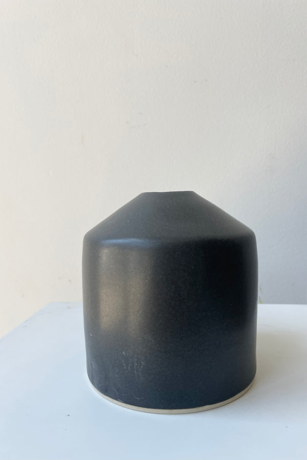 One small ceramic vase sits on a white surface in a white room. The vase is cylindrical and tapers off at the top to a narrow opening. The vase is black with a small ring of unglazed clay at the bottom. It is photographed straight on.