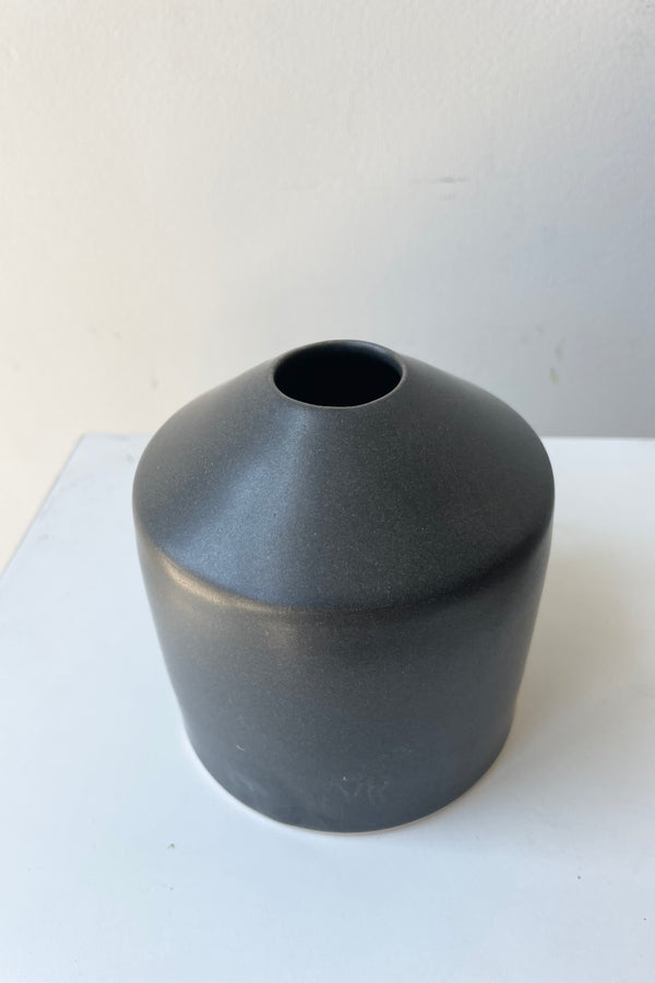 One small ceramic vase sits on a white surface in a white room. The vase is cylindrical and tapers off at the top to a narrow opening. The vase is black with a small ring of unglazed clay at the bottom. It is photographed from an overhead angle.