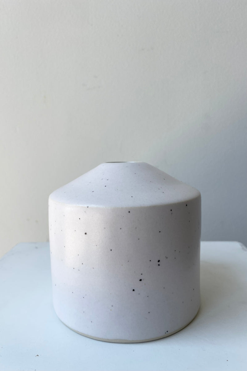 One small ceramic vase sits on a white surface in a white room. The vase is cylindrical and tapers off at the top to a narrow opening. The vase is white with black speckles and a small ring of unglazed clay at the bottom. It is photographed straight on.