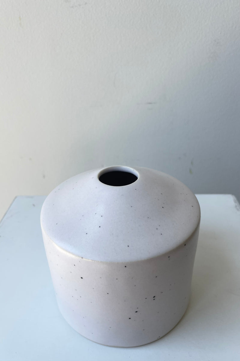 One small ceramic vase sits on a white surface in a white room. The vase is cylindrical and tapers off at the top to a narrow opening. The vase is white with black speckles and a small ring of unglazed clay at the bottom. It is photographed from an overhead angle.
