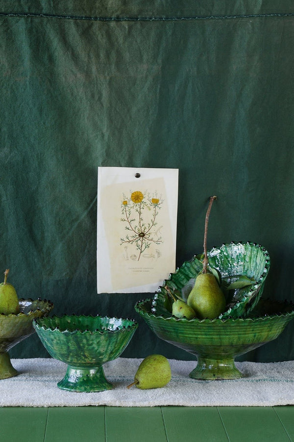 Various sized green tamegroute compote bowls against a green fabric background, a print with dandelions and pears.