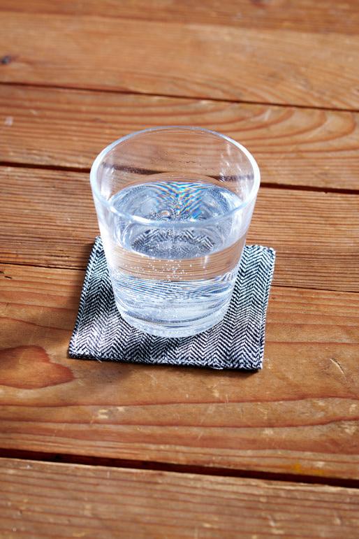 The Black Herringbone Coaster by Fog Linen under a glass with water sitting on a wood table. 