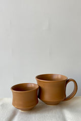 Photo of Little Fire Ceramics Hourglass cup and mug with a camel glaze on a white surface ahead of a white wall.