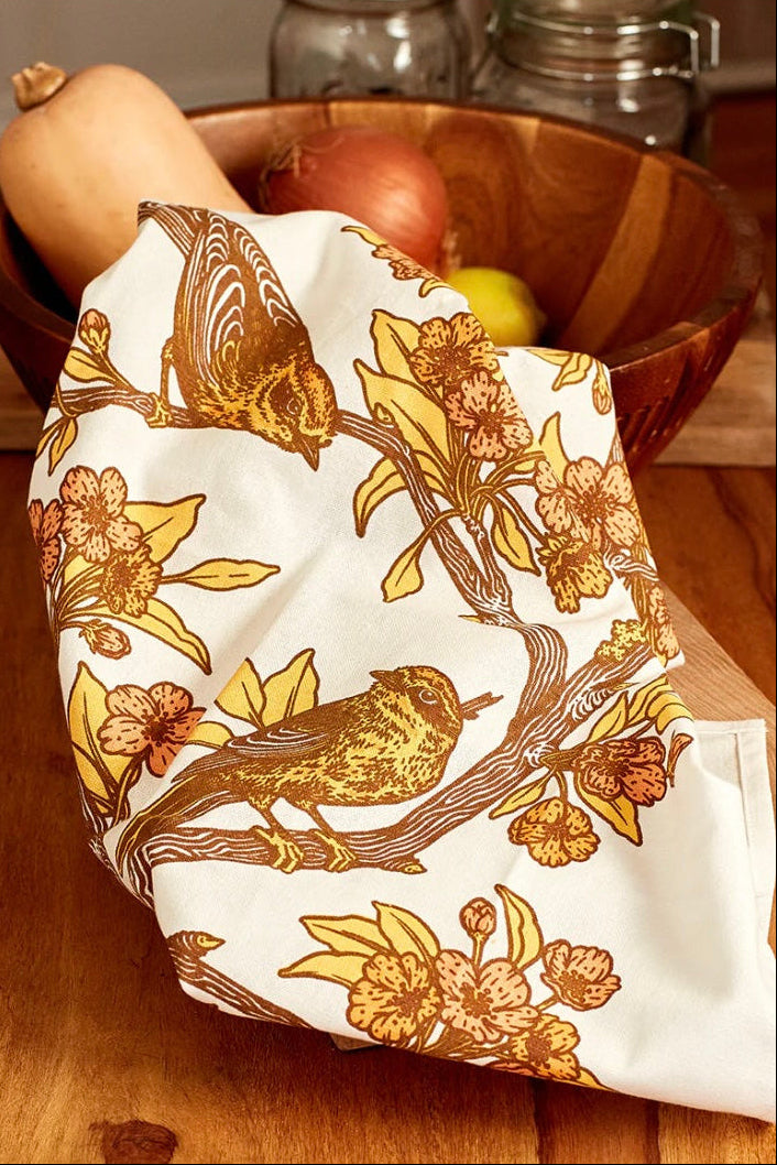 A pink and yellow screenprint of birds on branches on a white cotton tea towel on a wooden table ©Mustard Beetle