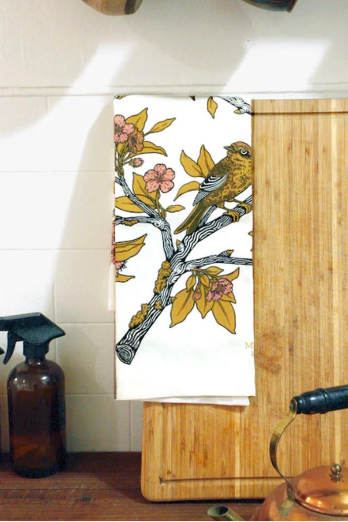 A pink and yellow screenprint of birds on branches on a white cotton tea towel with wood and white surface ©Mustard Beetle