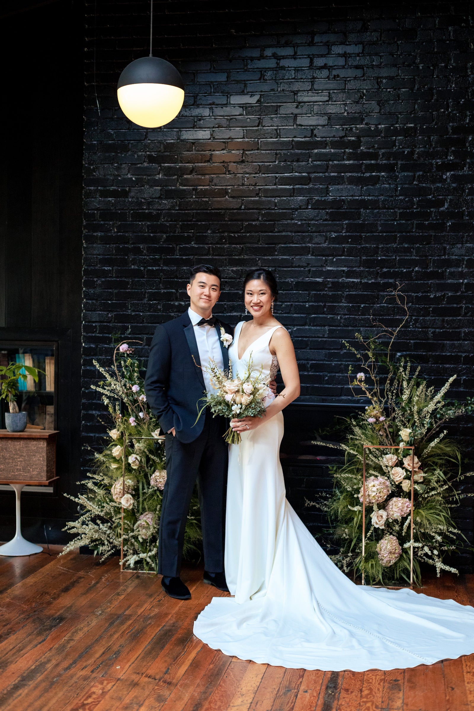 Tiffany and Andrew's wedding at the Moody Tongue Brewery. Bride and groom between two floor floral displays with textured foliage and larger cream floral, wild but structured. Bride holding her floral bouquet.  Black brick background wedding ceremony.  Floral by SPROUT HOME. Photograph by www.cageandaquarium.com