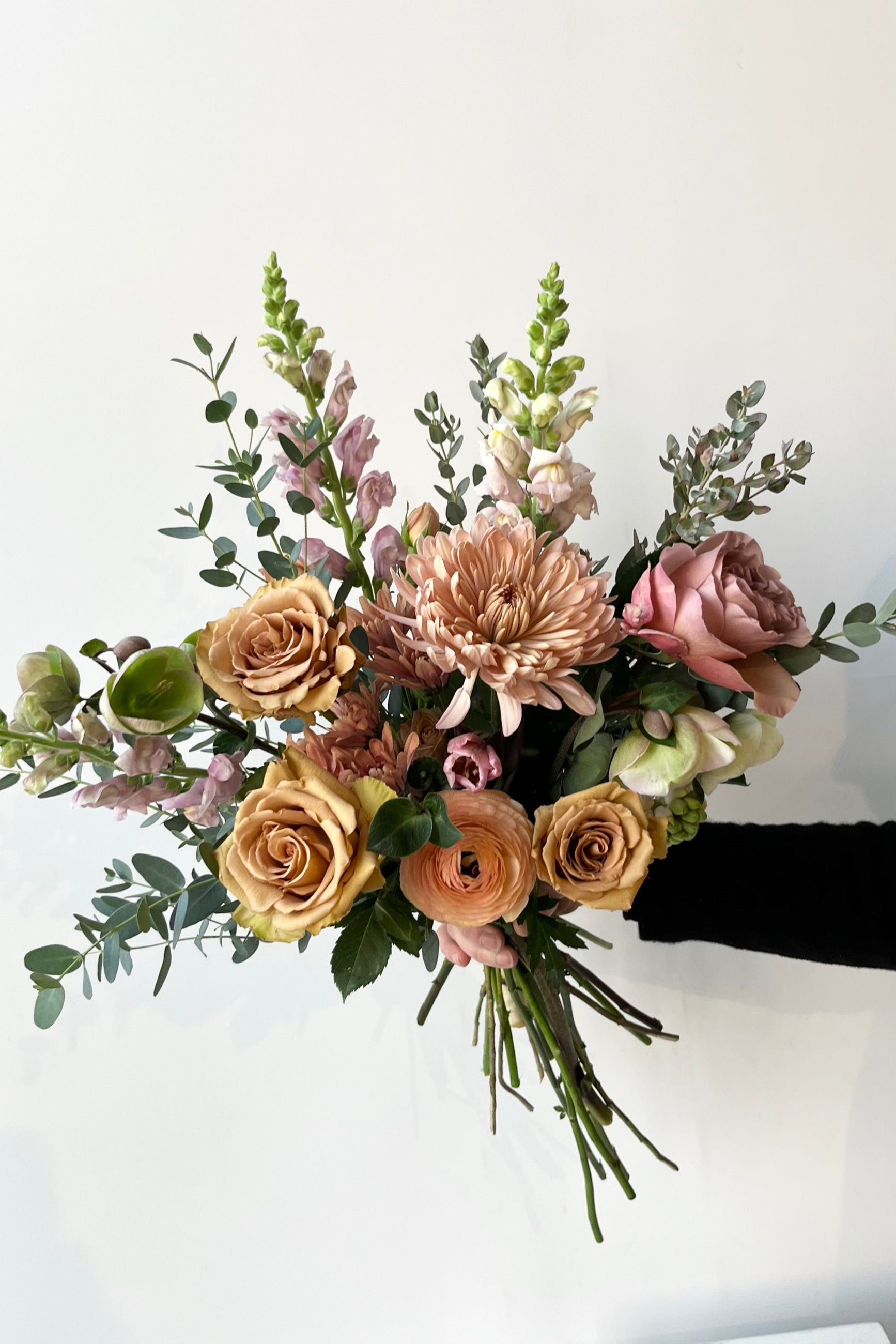 A bouquet held in front of a white wall with caramel and bronze colors roses and ranunculus and snapdragon with Hellbore in late winter ©Sprout Home #size_$100