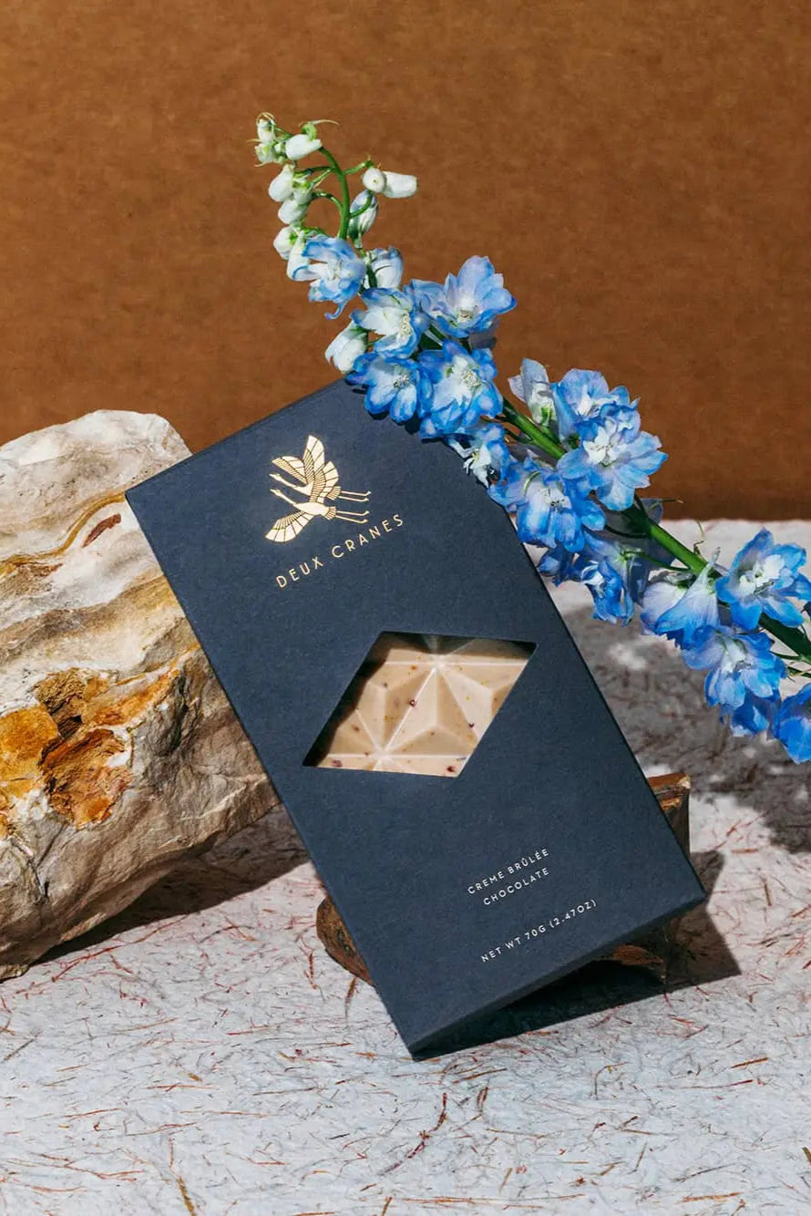 Photo of a Deux Cranes Creme Brulee Chocolate bar in a dark blue box with blue Delphinium flower in front of a stone background.