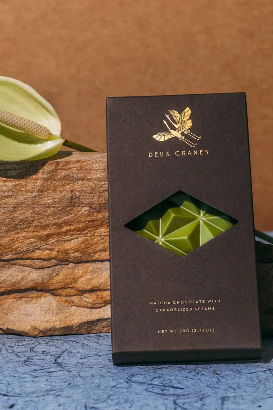 Photo of a packaged Deux Cranes Matcha Chocolate bar against wood and a brown wall.