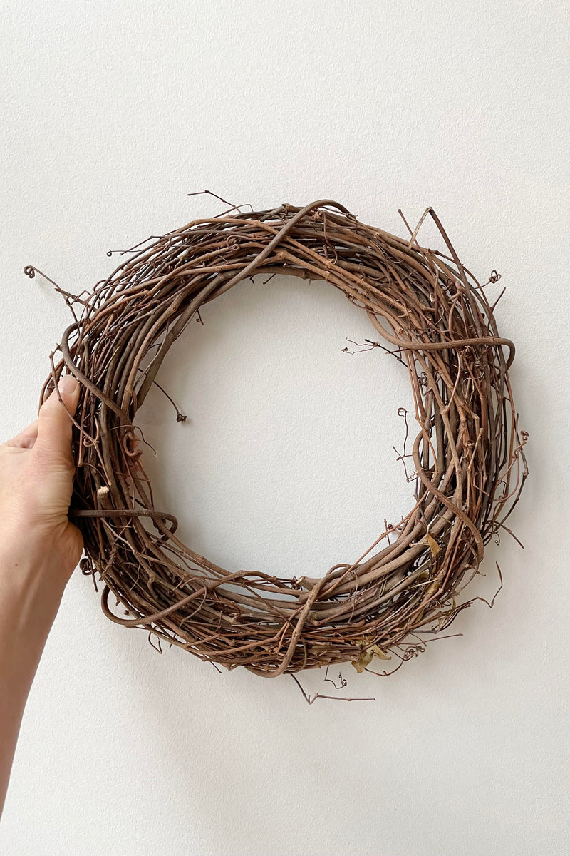 A hand holds a 12" wide grapevine wreath against a white wall.