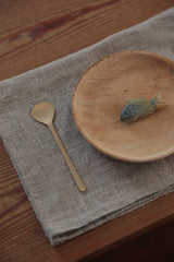 Brass dessert spoon by Fog Linen Work sitting on a linen cloth and next to a wood plate with a wood fish on it. 