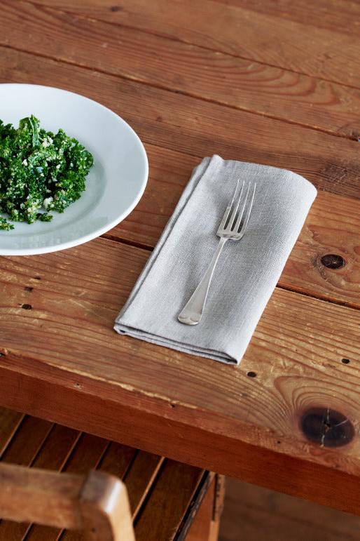 Fog Linen Work natural linen napkin folded with a form against a white plate of green on a wood table. 