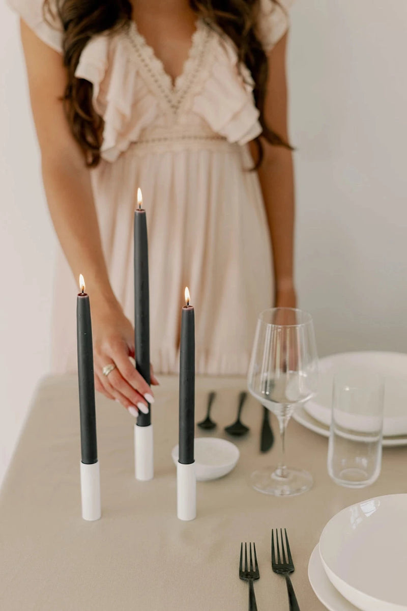 Mo & Co Charcoal beeswax taper candles shown lit and being placed in a table setting by a woman wearing a white dress.