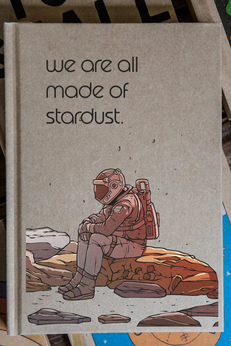 Notebook with an astronaut illustration and 'we are all made of stardust' text on a wooden surface. ©Glum Lot Press