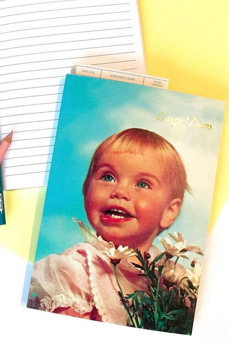 A vintage-style greeting card featuring a young blonde child with flowers, styled on a white surface with a lined notebook, blue and pink pencils, and yellow paper accents. ©Smitten Kitten