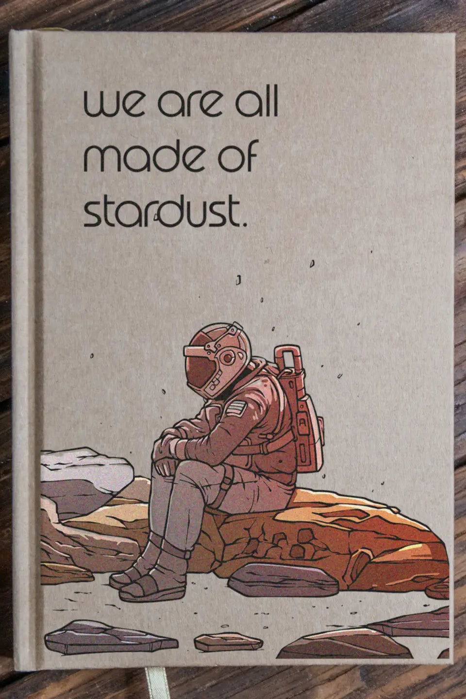 Notebook with an astronaut illustration and 'we are all made of stardust' text on a wooden surface. ©Glum Lot Press