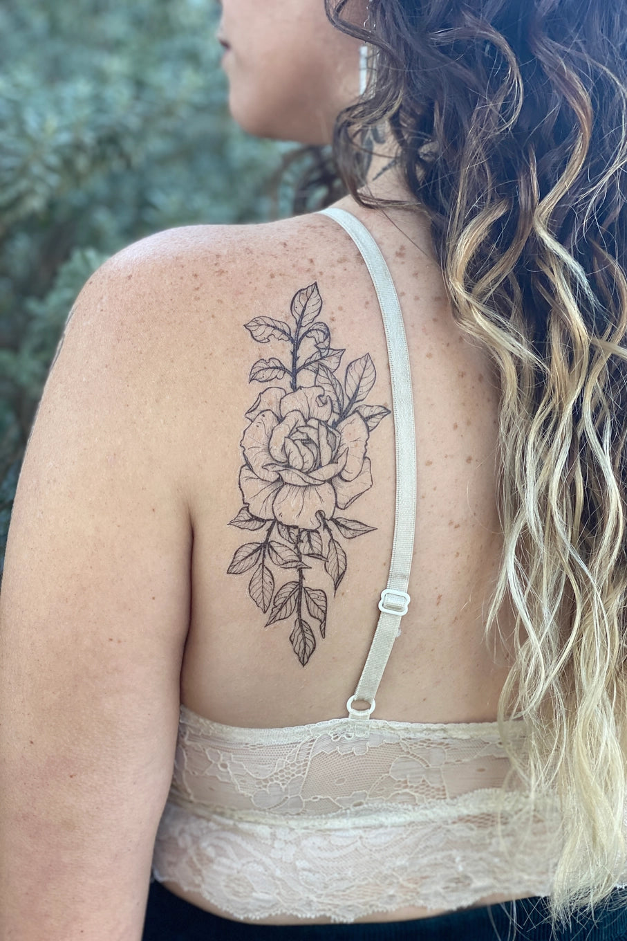 Woman with a Rose floral tattoo on her back, wearing a white lace top outdoors. ©Nature Tats