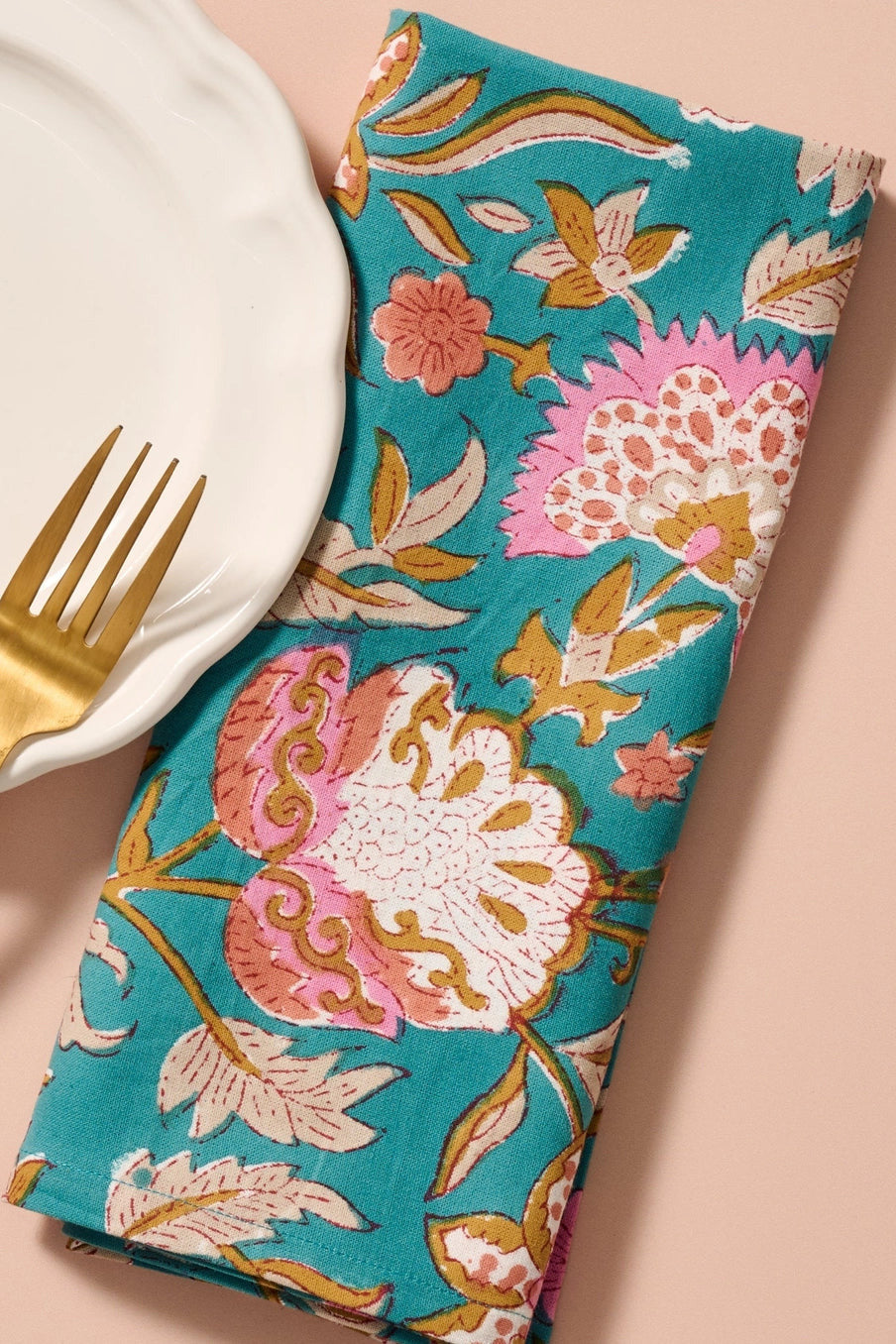 A folded Stabaka Block Print napkin with a pink, white, and gold floral pattern, styled on a peach background next to a white plate and a gold fork. ©Ten Thousand Villages