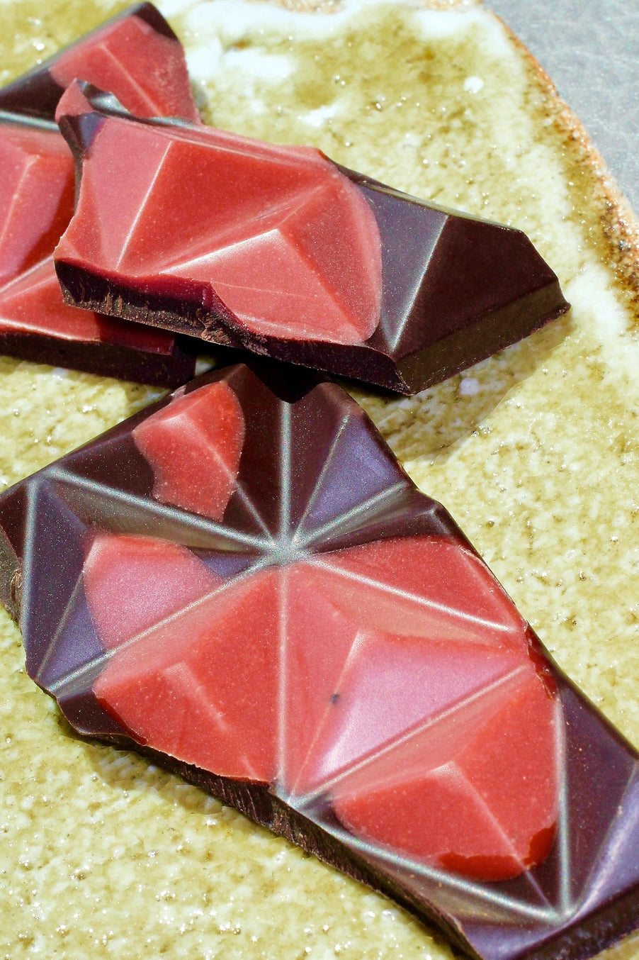 Geometrically shaped pieces of a dark chocolate and strawberries bar on a textured surface ©Deux Cranes
