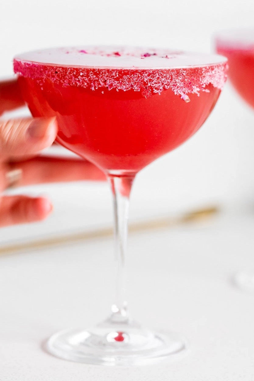 Red cocktail in a martini glass held by a hand on a white background ©Flouwer Co.