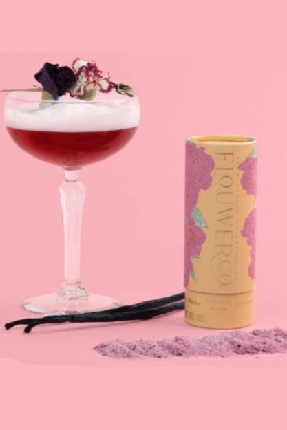 Cocktail with floral garnish next to a canister labeled 'Flowerco' on a pink background  ©Flouwer Co.