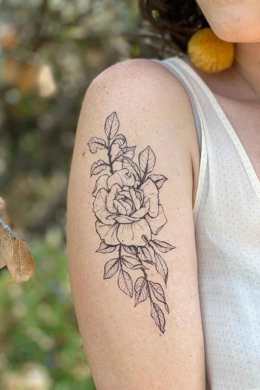Person with a floral Rose tattoo on their arm, standing outdoors with blurred natural background ©Nature Tats
