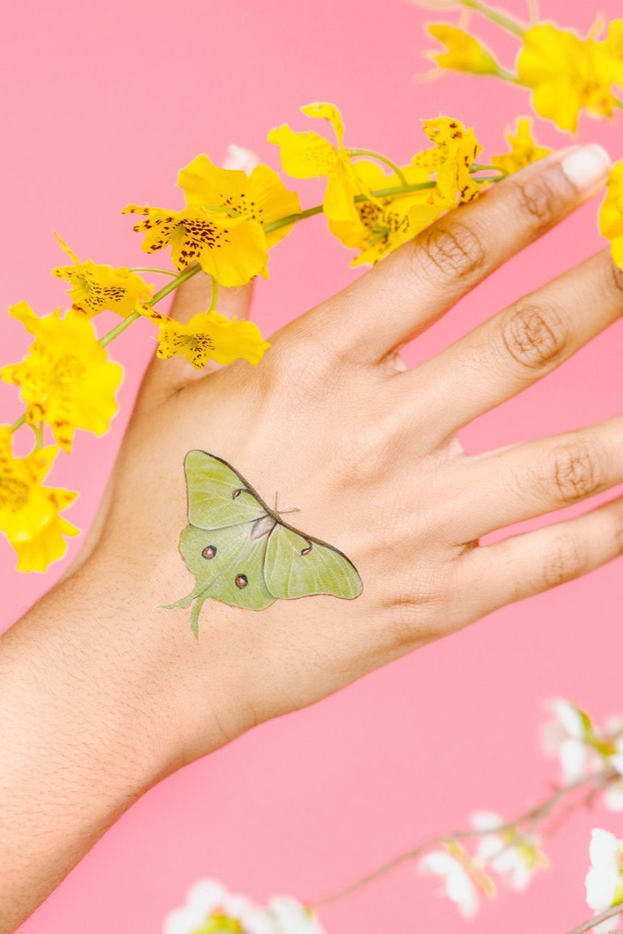Hand with a green butterfly tattoo from Tiny Butterfly Tattoo Tin holding yellow flowers against a pink background ©Tattly