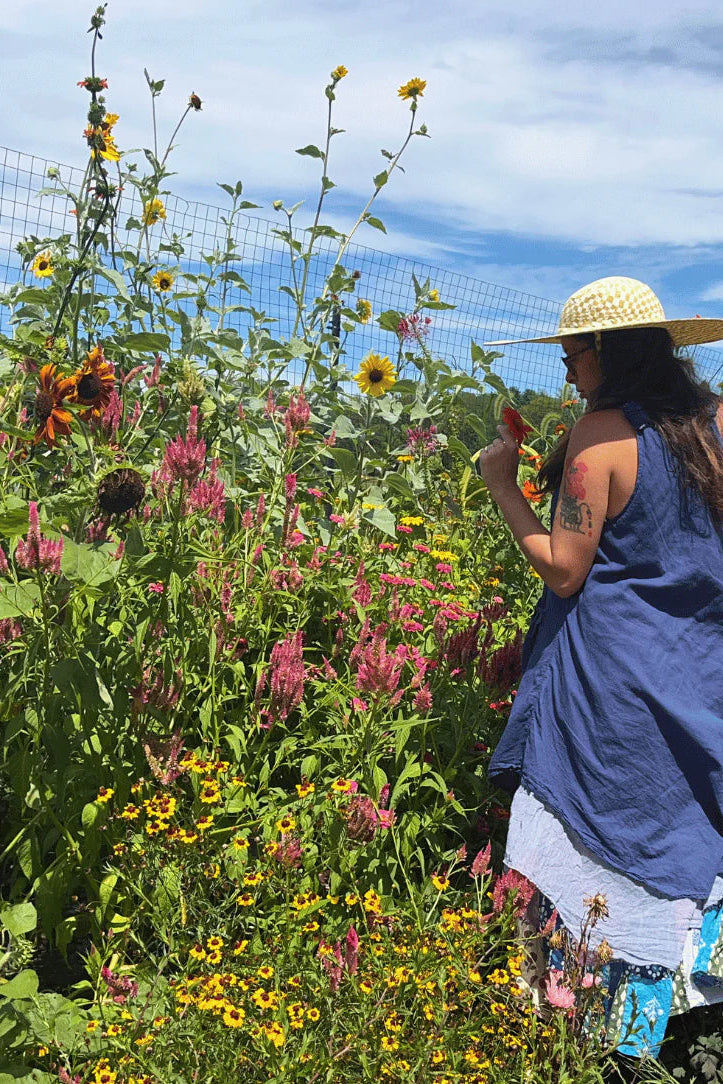 Woman in a cottage garden with flowers and plants, wearing a straw hat and blue dress. ©Hudson Valley Seed Co.
