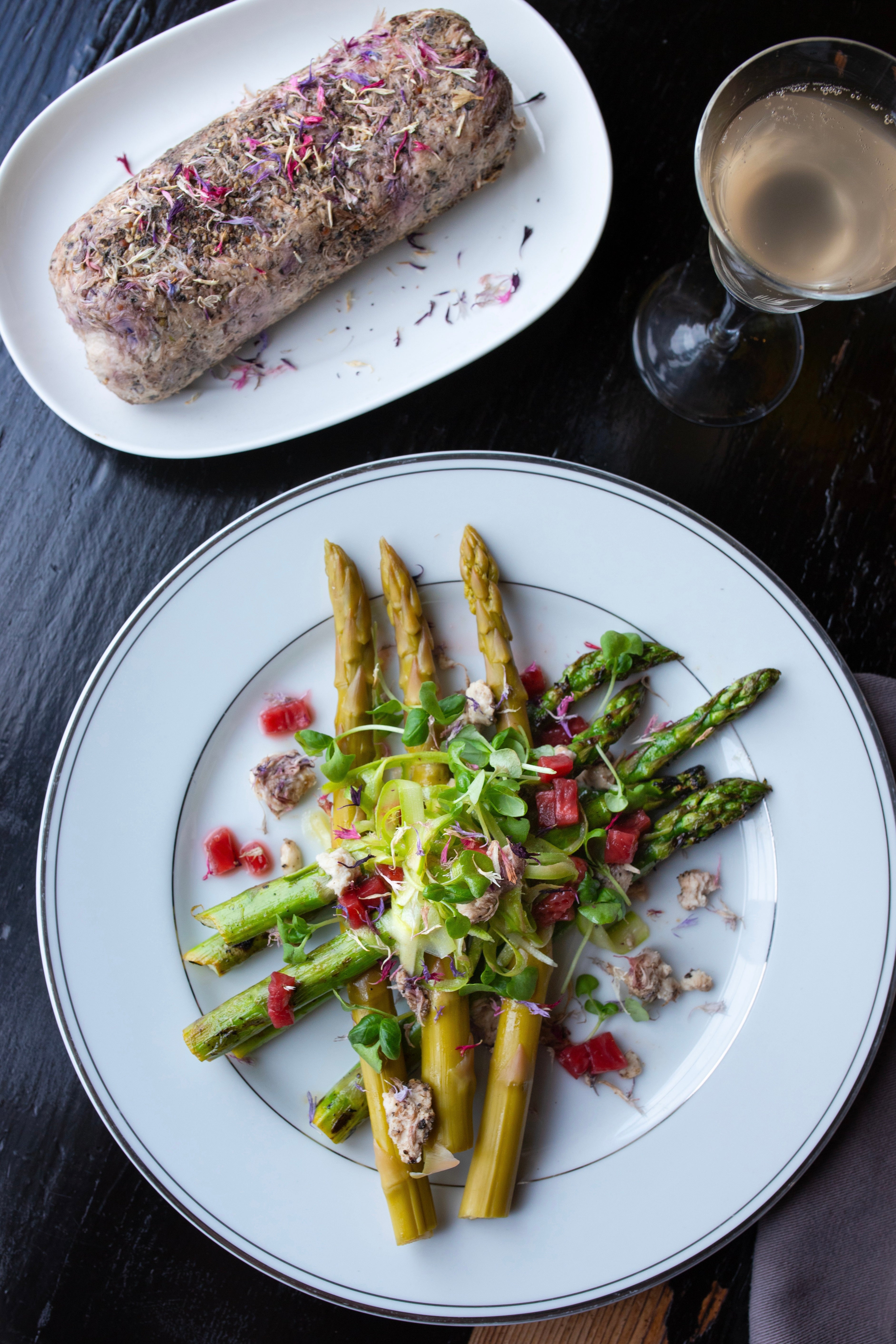 Photo of edible flower petals on an Asparagus salad plated on a round white plate on a black wood table. It is accompanied by a glass of wine and a bread plate, also topped with flowers. The photo is by chefs Mateja and Grills at Heritage Restaurant and Caviar Bar.