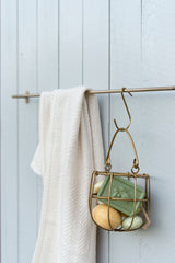 Small brass hook by Fog Linen Work in a bathroom on a brass towel holder holding  brass wire jar of soaps