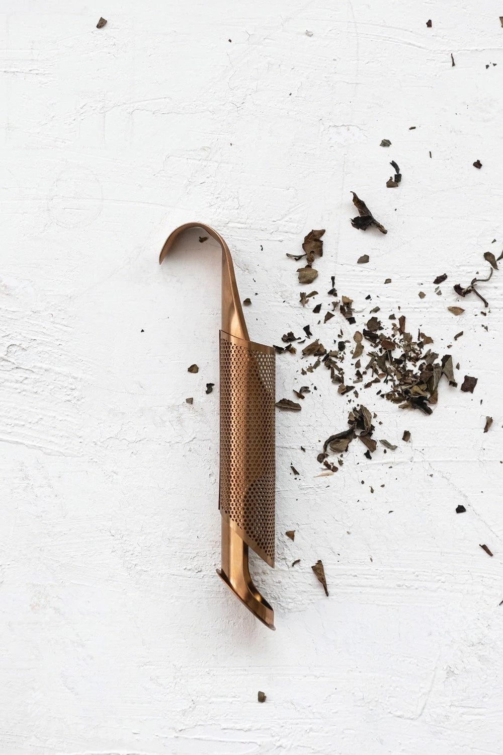 A rose gold colored tea infuser. The cylindrical infuser is on a white surface with scattered tea leavs ©Creative Co-Op