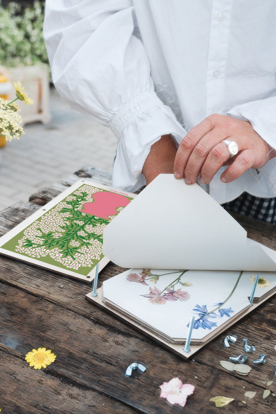 Person arranging pressed flowers in a Forget me Not Flower Press on a wooden surface with a blurred floral background. ©Studio Wald