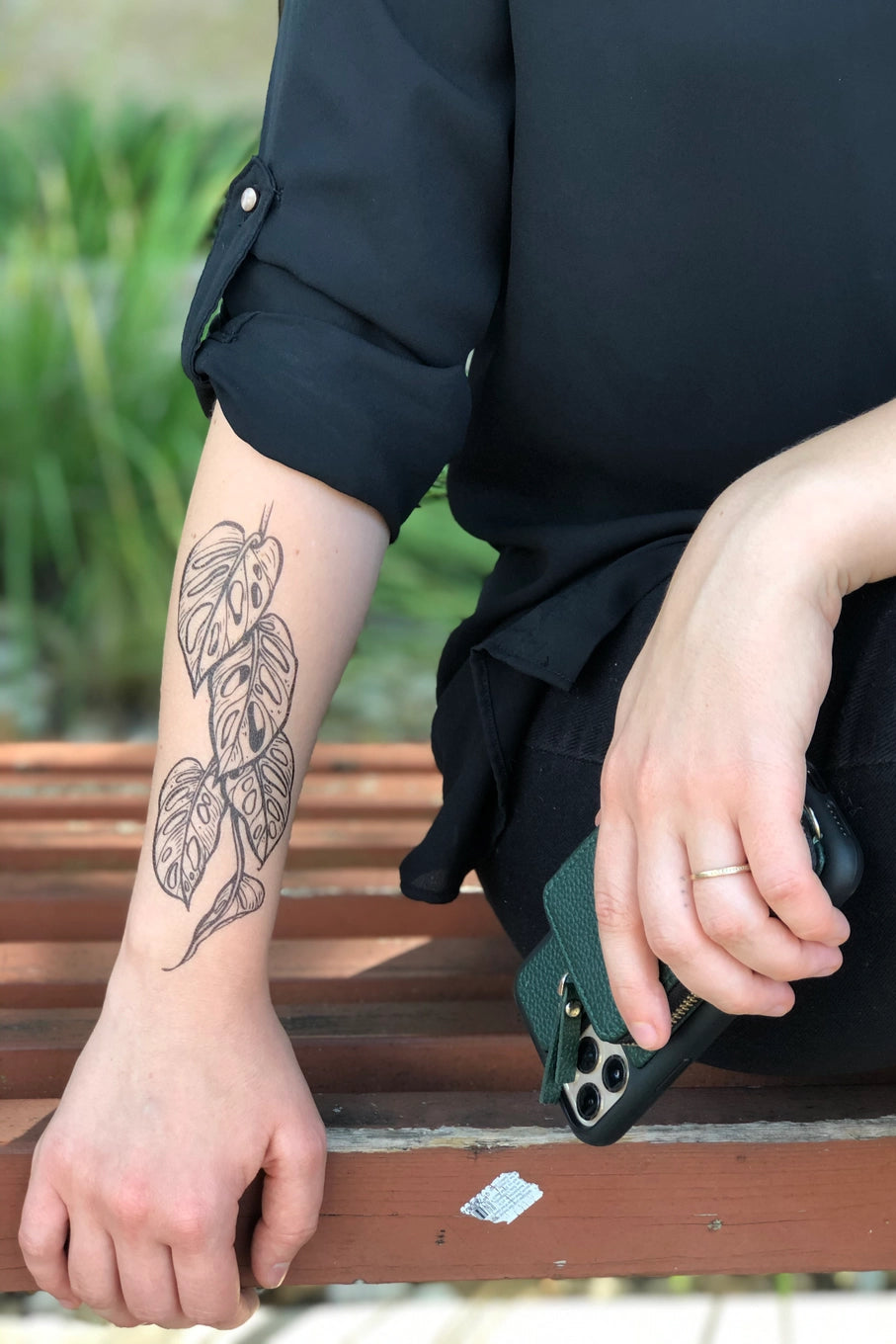 Person sitting on a bench with a tattoo on their arm, holding a phone case. ©Nature Tats