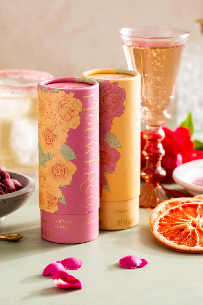Two floral-themed tins on a table with dried fruit, rose petals, and glasses in the background.  ©Flouwer Co.